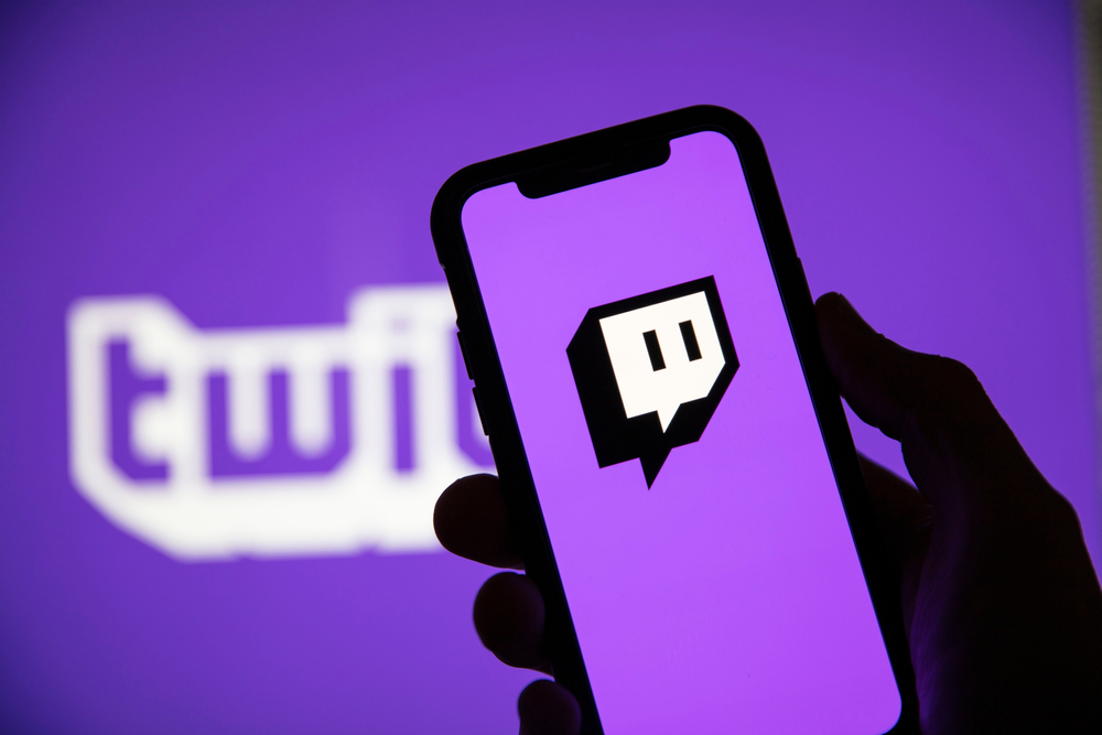 Hand holding smartphone showing Twitch icon with Twitch logo in background