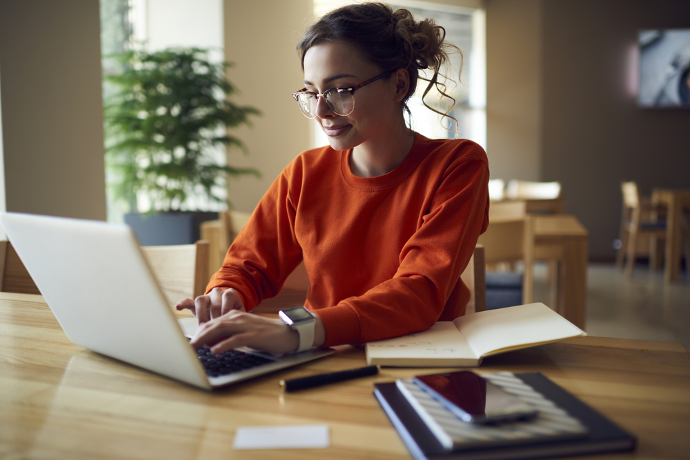 Young-female-copywriter-working-on-laptop-from-home-with-notes-on-desk-beside-her