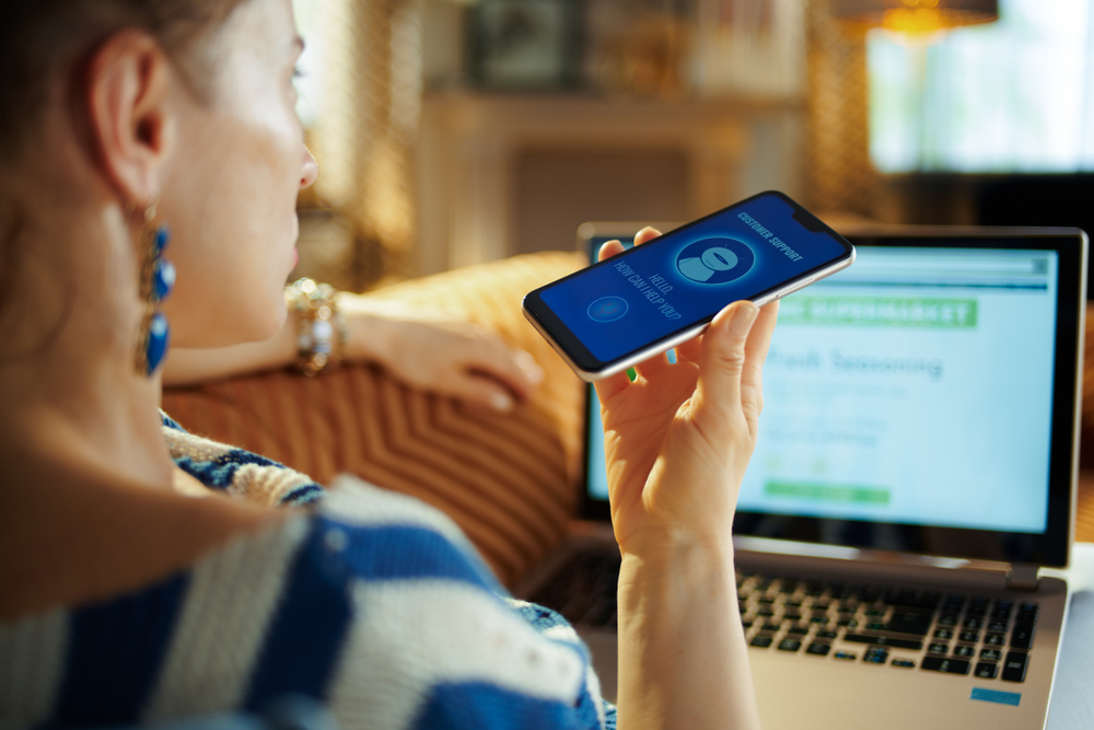 Woman-speaking-to-chatbot-on-her-phone-while-looking-at-her-laptop-computer.