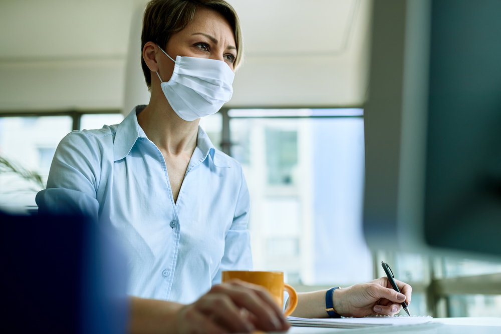 Woman-wearing-a-mask-working-on-a-computer-and-taking-notes