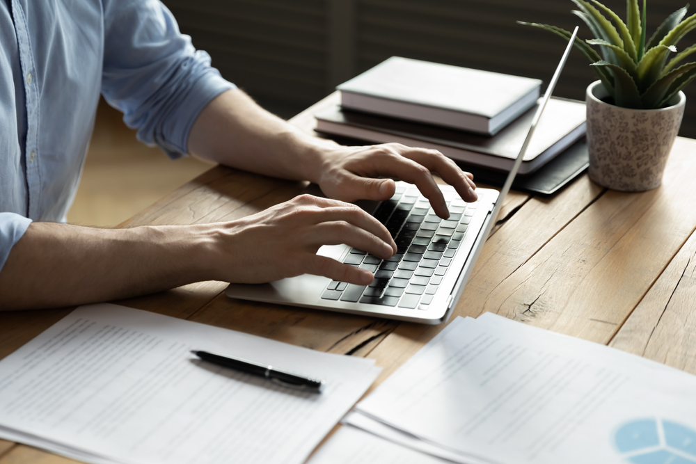 Man-typing-on-laptop-with-printed-notes-beside-him
