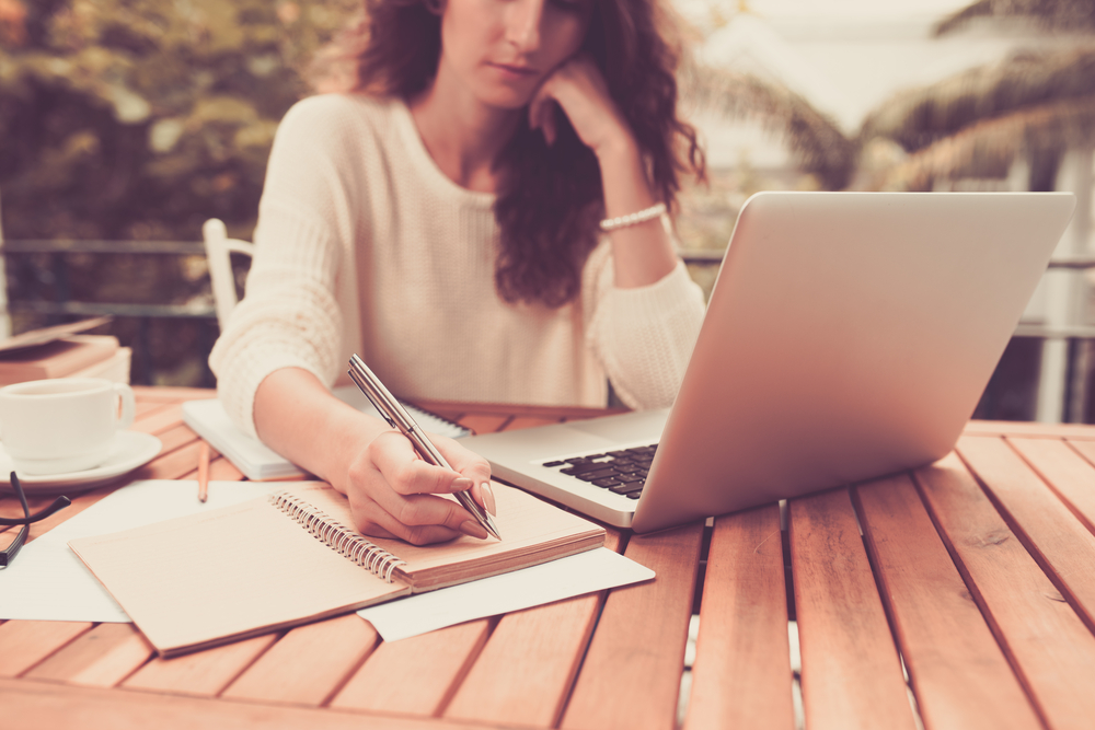 Woman-sitting-at-a-table-outdoors-with-laptop-and-notebook-with-pen