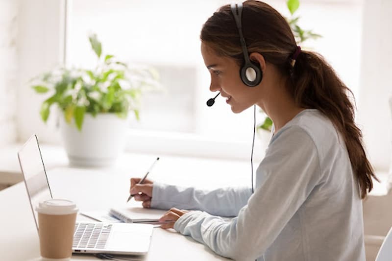 Young-woman-concentrating-writing-on-a-notepad-while-wearing-headphones-and-looking-at-laptop-screen
